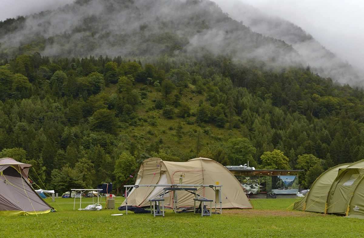 Plusieurs tentes beige et verte dressées dans prairie avec montagnes boisées brumeux à l'arrière-plan